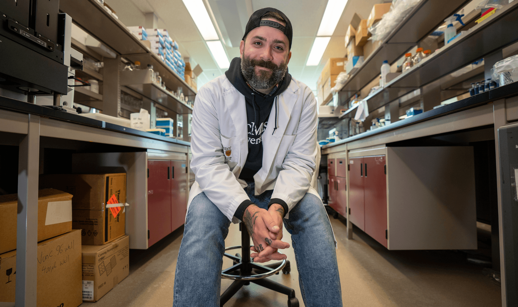 Jon Stokes, wearing a ball cap, a lab coat, a hoodie and jeans, sits in the middle of his lab, smiling down at the camera.